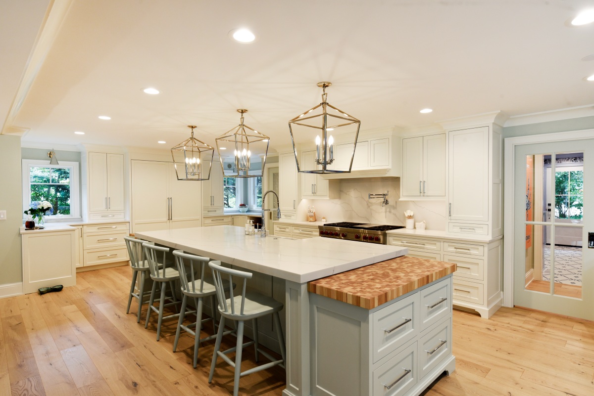 Bright white custom kitchen with large waterfall island, blue gray seating, butcher block prep ledge, glass lantern pendants, and wall of cabinetry around a professional range.