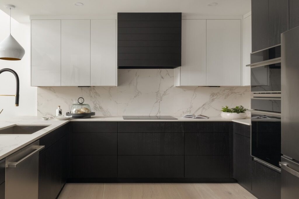 High end kitchen in Ludlow VT featuring two-tone cabinetry, a full-height marble-look backsplash, black wood base cabinets, and integrated appliances.